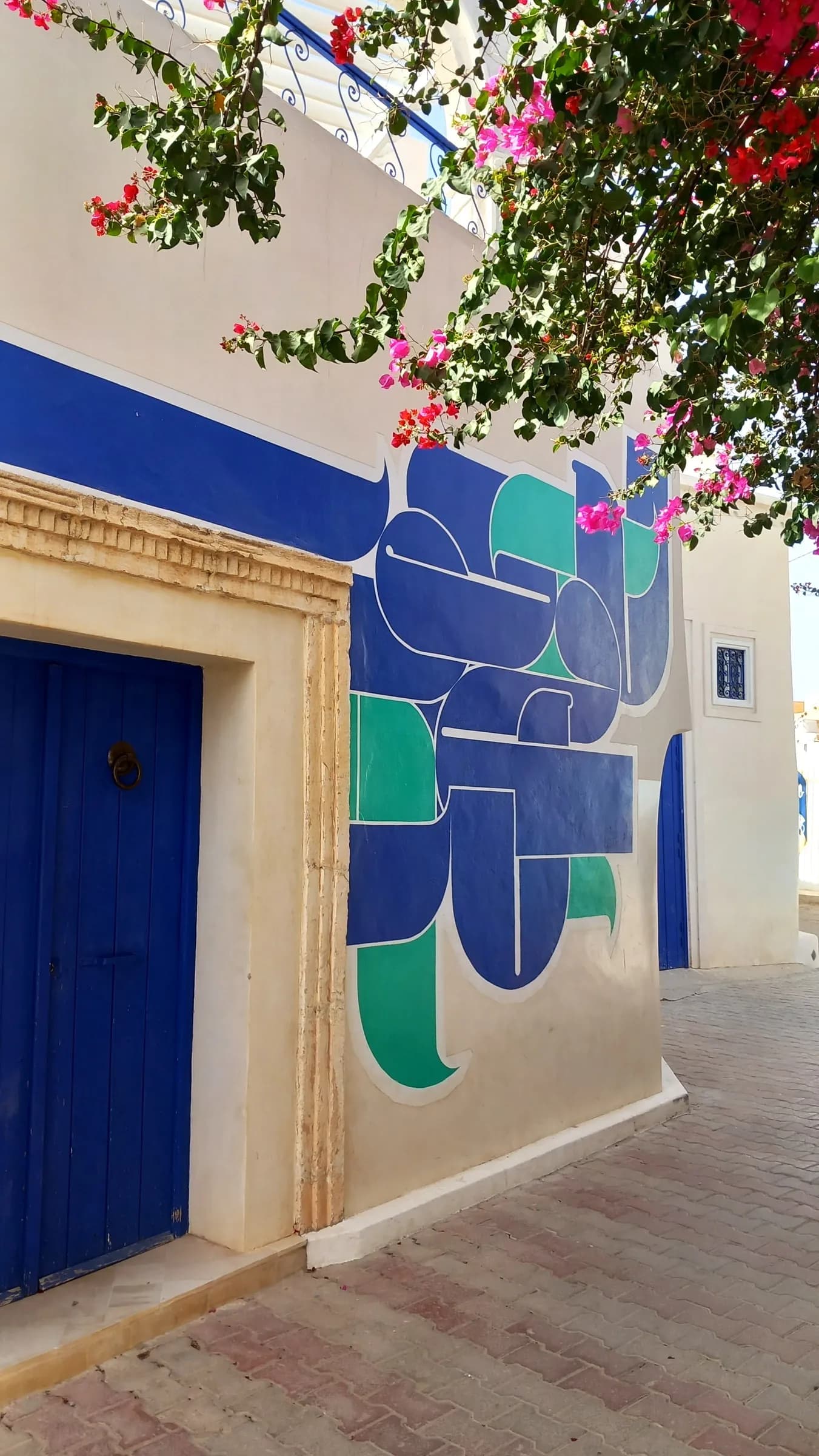 Un llamativo mural azul y verde en un muro encalado de Djerbahood, bajo las buganvillas